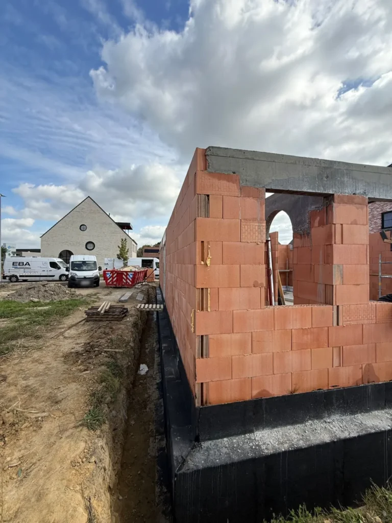 Zijgevel van een nieuwbouw in ruwbouw op een geïsoleerde funderingsmuur, met bestelwagens van EBA Construct op de werf